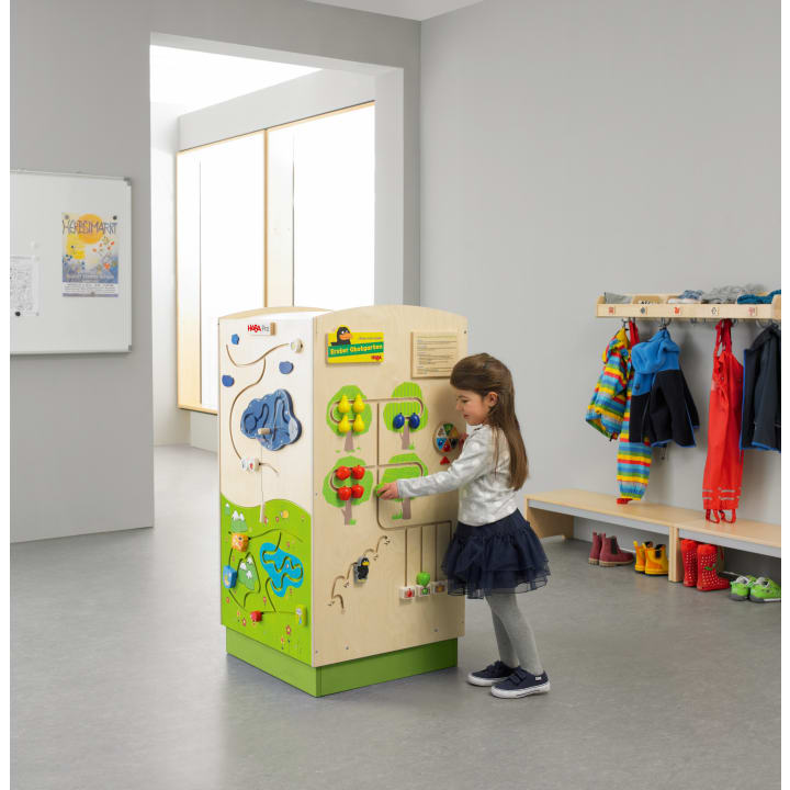 Ein junges Mädchen steht in einem Klassenzimmer und spielt mit einem großen Aktivitätswürfel aus Holz. Hinter ihr befinden sich Bänke, bunte Jacken, Schuhe und Ablagefächer. Der Raum hat einen grauen Boden, helle Wände und ein Fenster, das natürliches Licht hereinlässt.