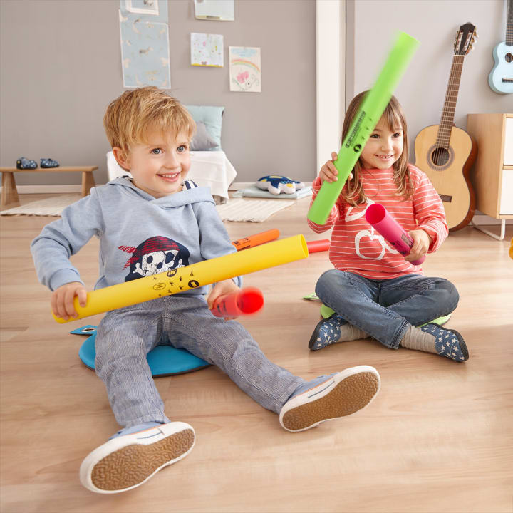 Zwei kleine Kinder sitzen in einem Raum auf dem Boden und halten jeweils bunte Plastikröhren in der Hand. Sie scheinen mit den Röhren zu spielen. Im Hintergrund sind Zeichnungen und eine Gitarre zu sehen.