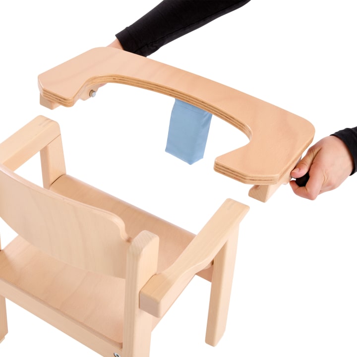 Eine Person ist dabei, ein Holztablett mit einem blauen Stoffband an einem Hochstuhl aus hellem Holz anzubringen oder abzunehmen. Der Hochstuhl und das Tablett sind vor einem weißen Hintergrund abgebildet.