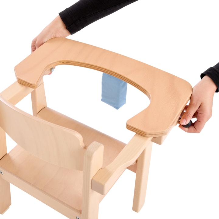 Eine Person befestigt ein gebogenes Holztablett an einem hellen Holzstuhl. Der Stuhl ist in der Mitte mit einem blauen Stoffband versehen, und die Hände der Person halten das Tablett an beiden Seiten.