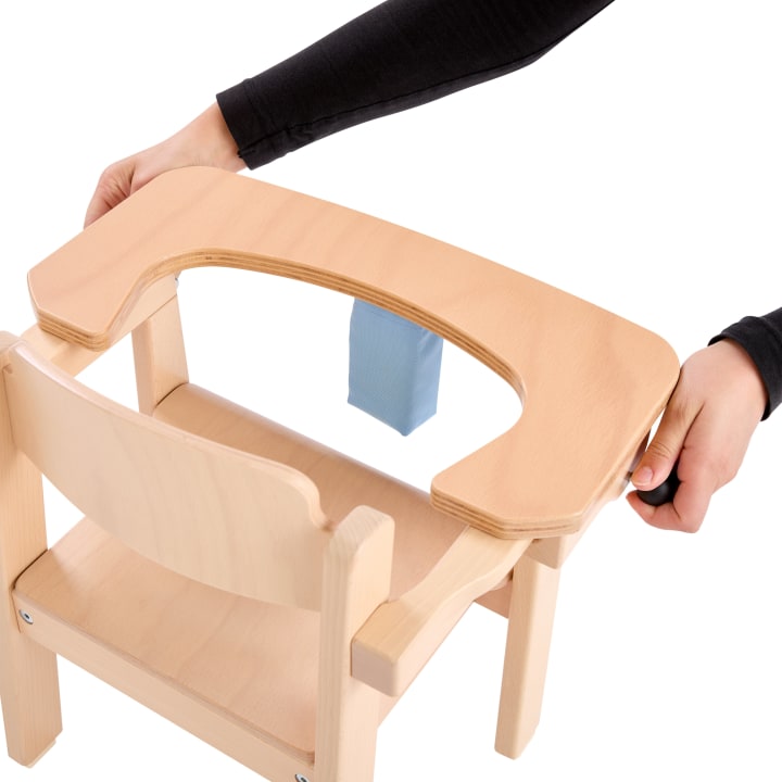 Eine Person befestigt ein Holztablett an einem hellen Holzstuhl mit einem blauen Gurt auf der Sitzfläche vor einem weißen Hintergrund.