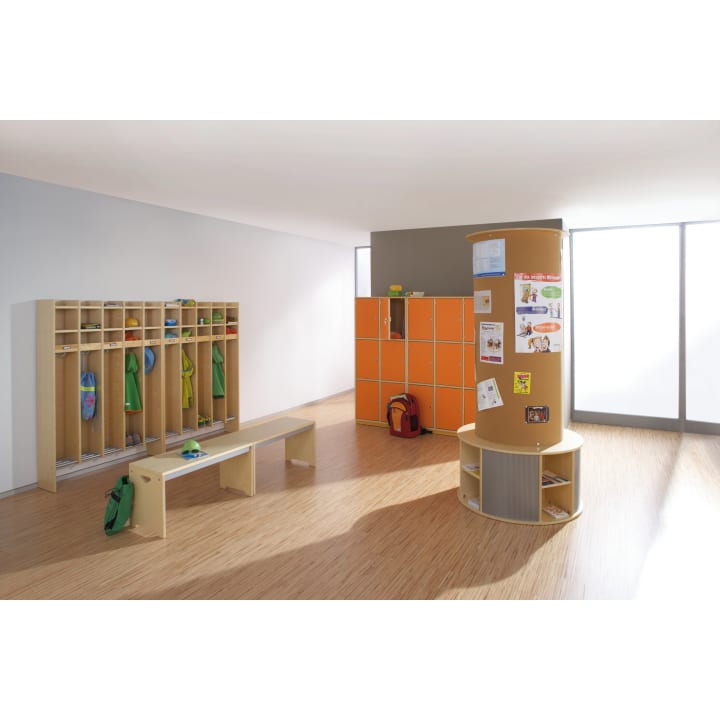Ein moderner, heller Raum mit hölzernen Fächern und orangefarbenen Schränken, einer Bank in der Mitte und einer zylindrischen Pinnwand mit verschiedenen Papieren und Zeichnungen. Rucksäcke und grüne Jacken hängen in den Fächern.