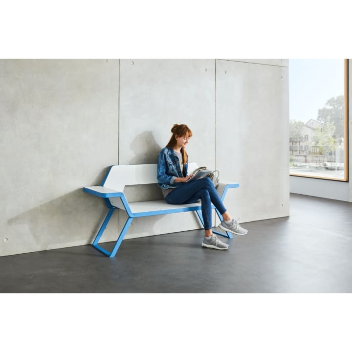 Eine Frau in Jeansjacke und Jeans sitzt auf einer modernen weiß-blauen Bank vor einer Betonwand und liest eine Zeitschrift. Auf der rechten Seite befindet sich ein großes Fenster, das natürliches Licht hereinlässt.
