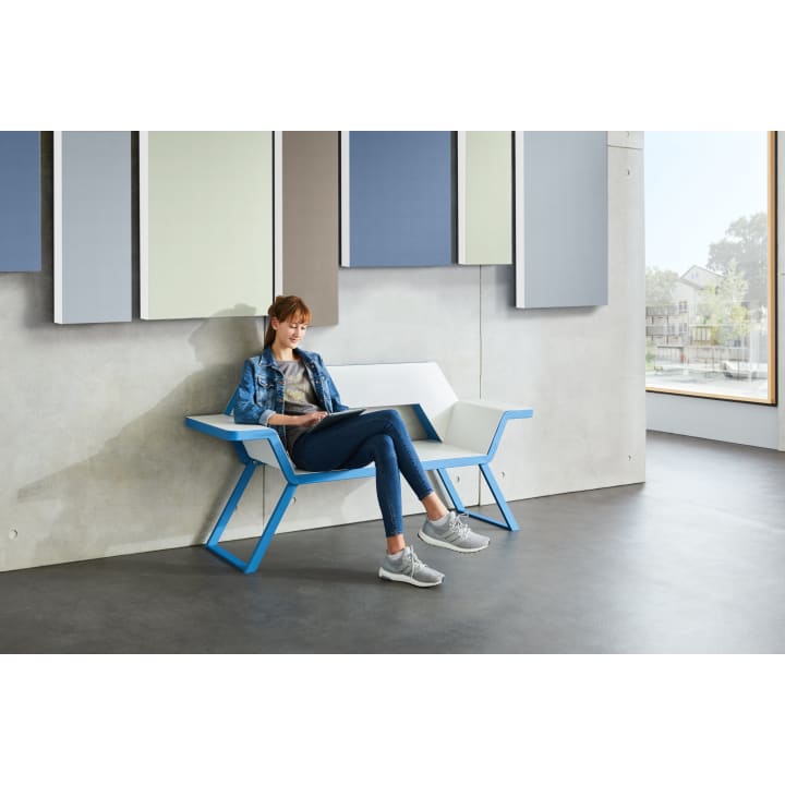 Eine Frau sitzt auf einer modernen weiß-blauen Bank vor einer Betonwand mit rechteckigen Akustikplatten. Sie schaut auf ein Tablet auf ihrem Schoß und trägt Jeans, ein graues T-Shirt, eine Jeansjacke und graue Turnschuhe.