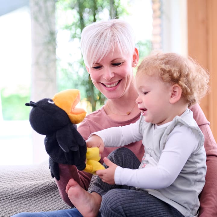 Ein Erwachsener mit kurzen weißen Haaren und ein kleines Kind sitzen zusammen auf einer Couch. Der Erwachsene hält eine schwarze Vogelhandpuppe, und das Kind interagiert mit ihr. Beide scheinen engagiert und auf die Puppe konzentriert zu sein.
