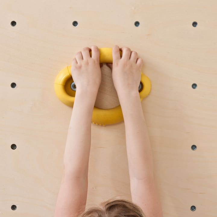 Die Hände eines Kindes halten einen gelben Klettergriff, der an einer hellen hölzernen Kletterwand mit gleichmäßig verteilten Löchern befestigt ist.
