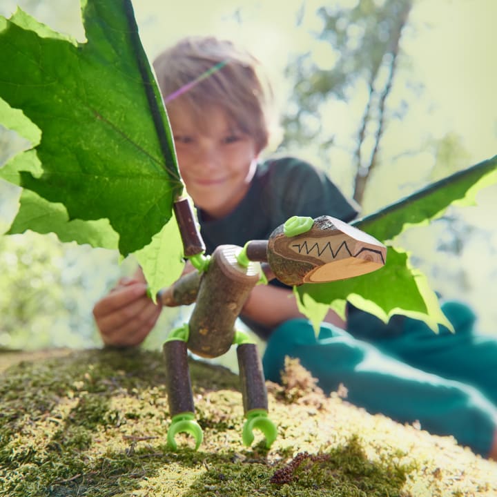 Ein Kind hockt im Freien hinter einer handgefertigten Drachenfigur aus Stöcken, Blättern und Spielzeuggelenken, die auf moosigem Boden steht, während das Sonnenlicht durch die Bäume im Hintergrund fällt.