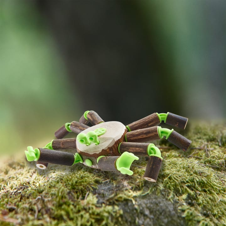 Eine kleine Spielzeugspinne aus grünen Plastiksteckern und braunen Holzstäben sitzt auf Moos, wobei der unscharfe grüne Hintergrund eine natürliche Umgebung im Freien andeutet.