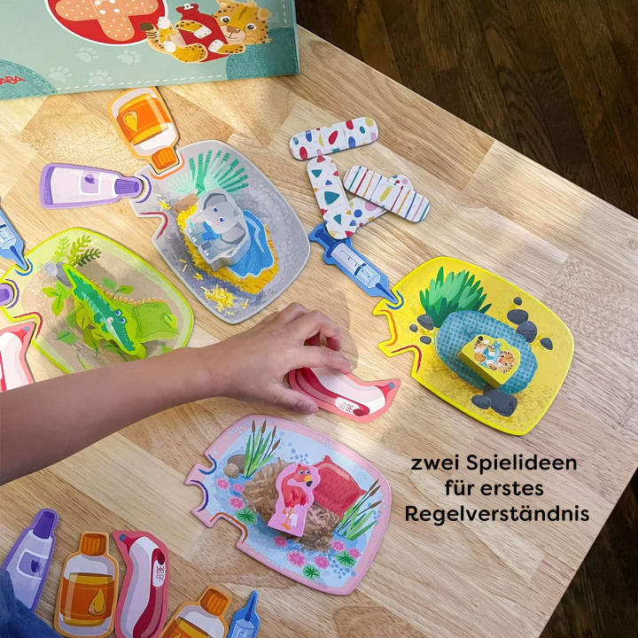 Die Hand eines Kindes setzt auf einem Holztisch bunte Puzzleteile mit Tiermotiven zusammen. Andere Puzzleteile, auf denen Flaschen und Tiere abgebildet sind, liegen verstreut herum. Der deutsche Text lautet: Zwei Spielideen für erstes Regelverständnis.
