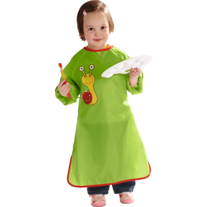 Ein kleines Kind, das eine grüne Schürze mit Schneckenmotiv trägt, einen Pinsel in der einen und eine weiße Palette in der anderen Hand hält, steht und schaut nach vorne.