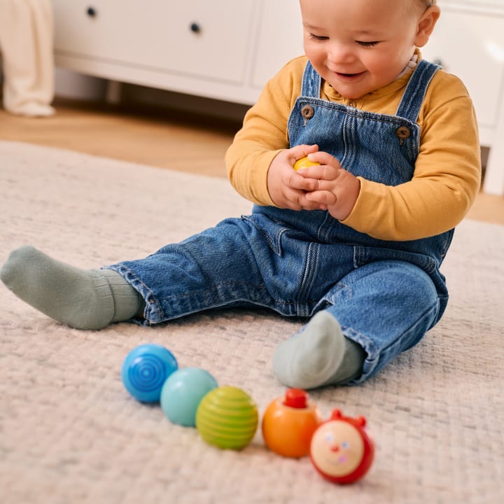 Ein Baby in Jeans-Overall und gelbem Hemd sitzt auf einem Teppichboden, hält einen gelben Spielzeugball in der Hand und lächelt, während vor ihm mehrere bunte Spielzeugbälle aufgereiht sind.