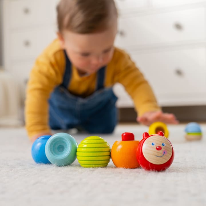 Ein kleines Kind in einem gelben Hemd und einem blauen Overall krabbelt auf einem Teppichboden und greift nach buntem Holzspielzeug, darunter ein rotes Spielzeug mit einem lächelnden Gesicht, vor einer verschwommenen Kommode im Hintergrund.
