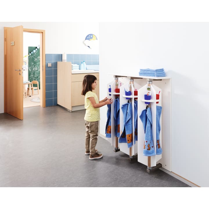 Ein kleines Kind steht vor einem Regal mit blauen Handtüchern und Flaschen und greift nach einem beschrifteten Haken. Die Szene spielt in einem hellen, aufgeräumten Klassenzimmer oder in einer Kindertagesstätte mit einer offenen Tür zu einem anderen Raum.