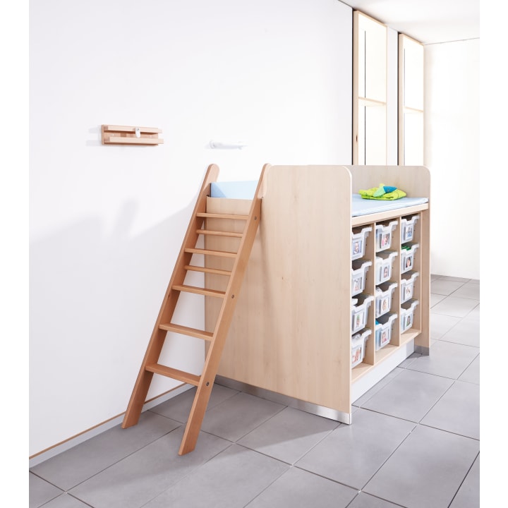 Eine Wickelkommode aus hellem Holz mit einer Leiter, einer Wickelauflage, Babyzubehör oben und 12 beschrifteten Aufbewahrungsbehältern unten, auf einem gefliesten Boden vor einer weißen Wand.