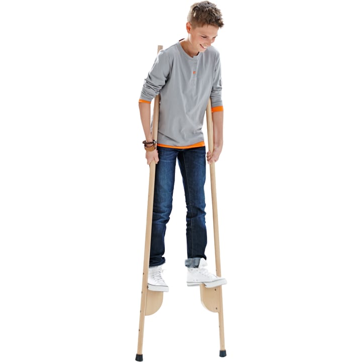 Ein Jugendlicher in einem grauen, langärmeligen Hemd und blauen Jeans steht auf Holzstelzen. Er lächelt und schaut nach unten, während er auf den Stelzen balanciert. Der Hintergrund ist weiß.