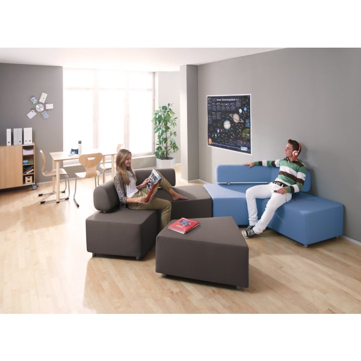 Zwei junge Menschen sitzen auf modernen, modularen Sofas in einem hellen Raum mit Holzboden. Einer liest eine Zeitschrift, während der andere Kopfhörer trägt. Im Hintergrund stehen ein Schreibtisch, Stühle, Bücherregale und eine Pflanze.