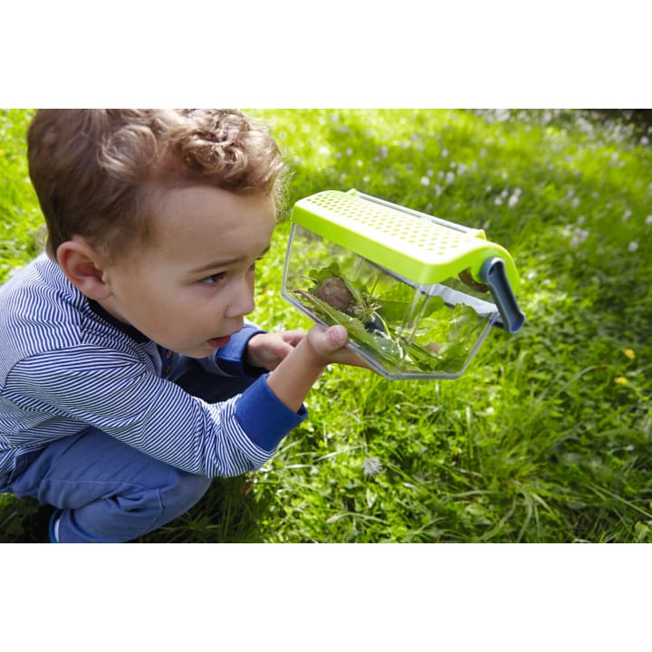 Ein kleines Kind hockt im Gras und hält einen durchsichtigen Plastikbehälter mit grünem Deckel, in dem es eine Schnecke und Blätter beobachtet.