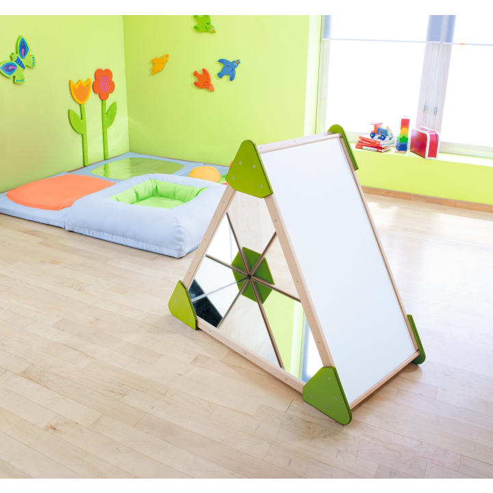 Ein Kinderspielzimmer mit grünen Wänden, weichen Spielmatten, Wanddekorationen mit Blumen und Schmetterlingen und einer dreieckigen sensorischen Spielstruktur mit Spiegeln und weißen Tafeln auf einem Holzboden.