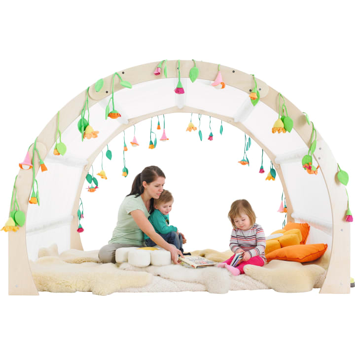 Ein Erwachsener und zwei kleine Kinder sitzen auf Kissen und Decken unter einem mit Stoffblumen und Blättern verzierten Holzbogen. Der Erwachsene liest einem Kind vor, während das andere in einem Buch blättert. Die Umgebung wirkt gemütlich und verspielt.