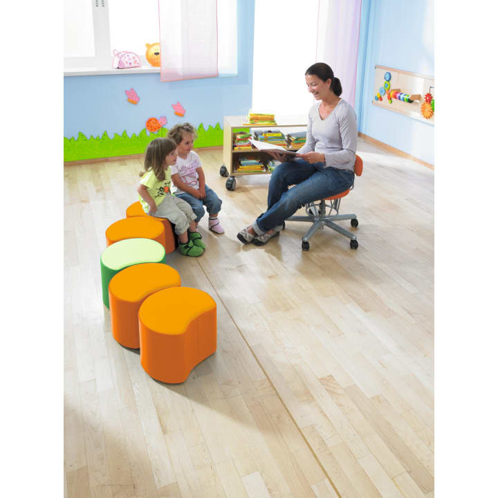 Eine Frau sitzt auf einem Stuhl und liest zwei kleinen Kindern vor, die auf orangefarbenen und grünen Hockern in einem hellen Klassenzimmer mit Holzfußboden und pädagogischen Dekorationen an den Wänden sitzen.