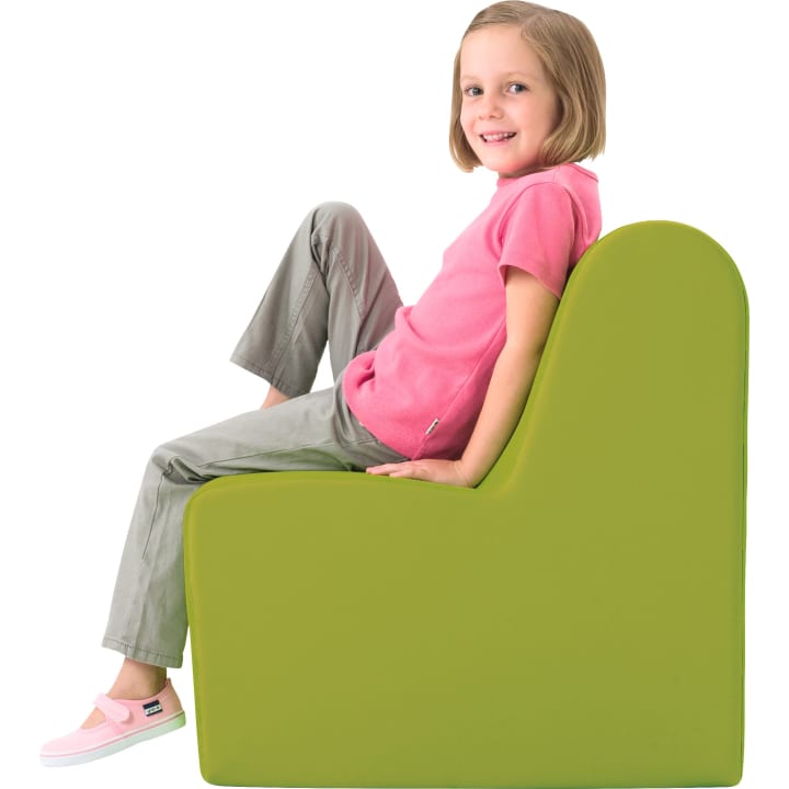 Ein kleines Kind mit kurzen blonden Haaren, das ein rosa Hemd, eine graue Hose und rosa Schuhe trägt, sitzt entspannt und lächelnd auf einem großen, grünen, L-förmigen Schaumstoffstuhl. Der Hintergrund ist weiß.