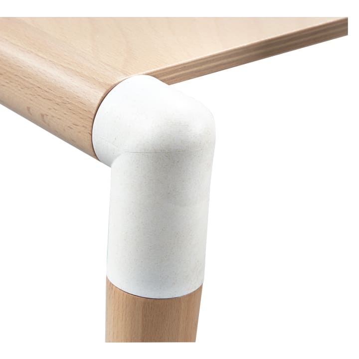 Nahaufnahme einer hölzernen Tischecke, die ein abgerundetes weißes Verbindungsstück zeigt, das die Tischplatte mit dem Bein verbindet, beide aus hellem Holz. Das Verbindungsstück scheint aus einem anderen Material als das Holz zu bestehen.