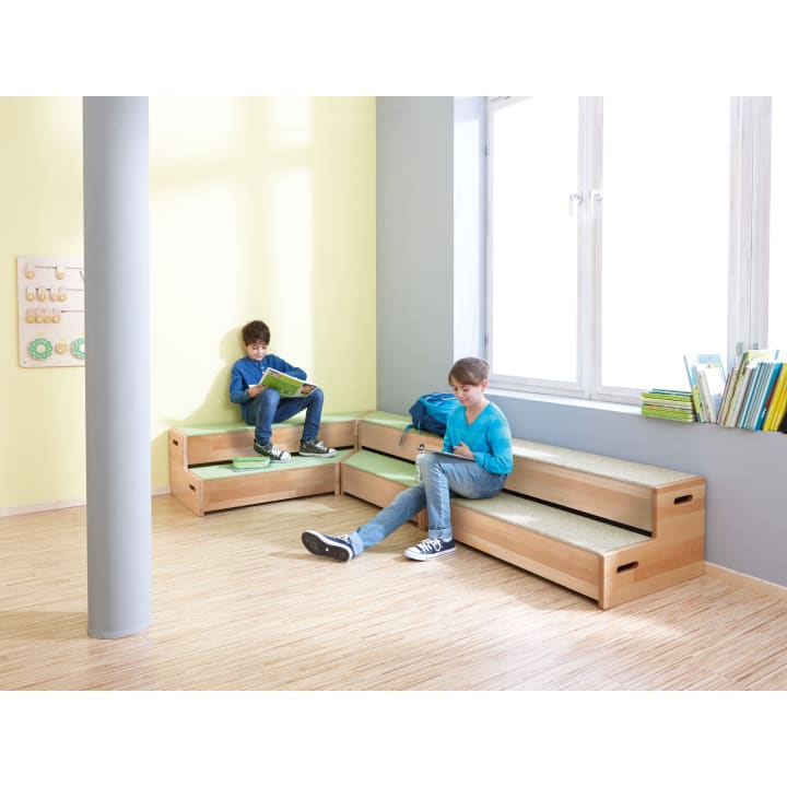 Zwei Jungen sitzen auf einer Holzbank in der Ecke eines hellen Raums mit hellem Holzboden und großen Fenstern. Ein Junge liest in einem Buch, der andere schreibt in ein Notizbuch. In der Nähe sind mehrere Bücher gestapelt.
