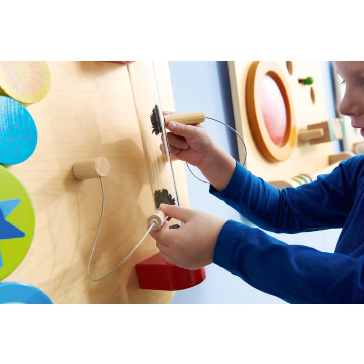 Ein Kind in einem blauen langärmeligen Hemd interagiert mit einem an der Wand befestigten Aktivitätsbrett aus Holz, indem es mit beiden Händen einen an einem Draht befestigten Griff durch ein Labyrinth mit Zahnrädern bewegt.