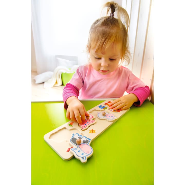Ein kleines Kind mit blonden Haaren in einem Pferdeschwanz und einem rosafarbenen Hemd sitzt an einem grünen Tisch und legt ein tierförmiges Puzzleteil in ein hölzernes Puzzlebrett.