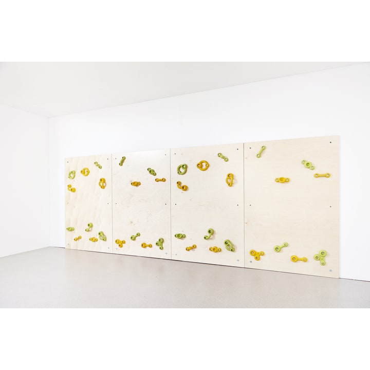 Eine helle Indoor-Kletterwand aus Holz mit vier Paneelen, auf denen jeweils grüne und gelbe Klettergriffe angebracht sind, an einer weißen Wand in einem hellen, leeren Raum mit grauem Boden.