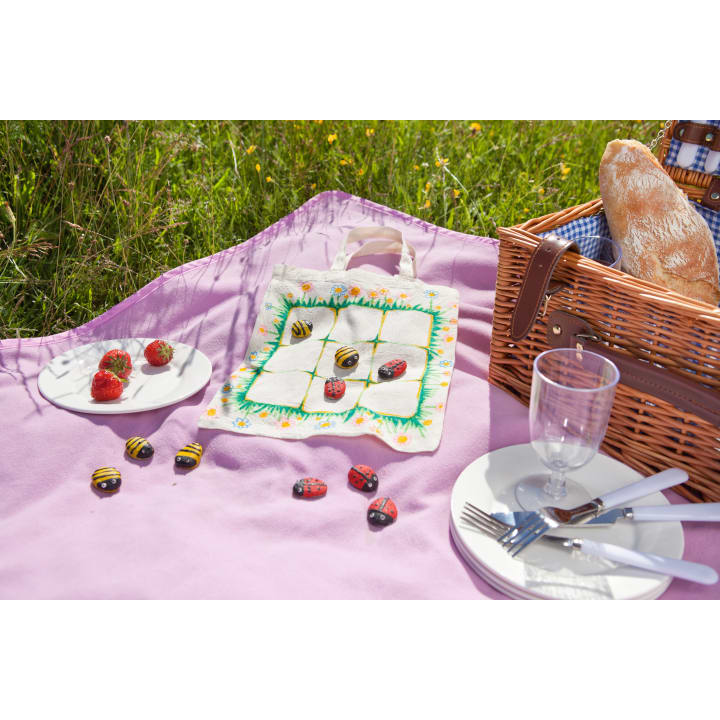 Ein Picknick auf einer Lavendeldecke mit einem Weidenkorb, Brot, Tellern, Besteck, Gläsern, Erdbeeren auf einem Teller und einer Serviette, die als Tic-Tac-Toe-Brett mit aufgemalten Steinbienen und Marienkäfern dient.