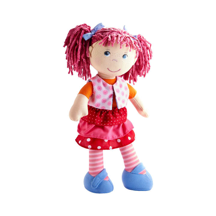 Eine weiche Stoffpuppe mit rosa Garnhaaren, die zu Zöpfen gestylt sind. Sie trägt eine rosa gemusterte Weste, einen roten Rock mit Tupfen, ein orangefarbenes Hemd, gestreifte rosa Leggings und blaue Schuhe. Die Puppe hat einen neutralen Gesichtsausdruck.