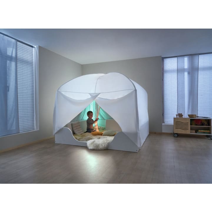 Ein Kind sitzt in einem weißen Innenzelt mit lichtdurchlässigen Vorhängen, umgeben von Kissen und einem flauschigen Teppich, in einem minimalistischen Raum mit großen Fenstern und Holzfußboden.
