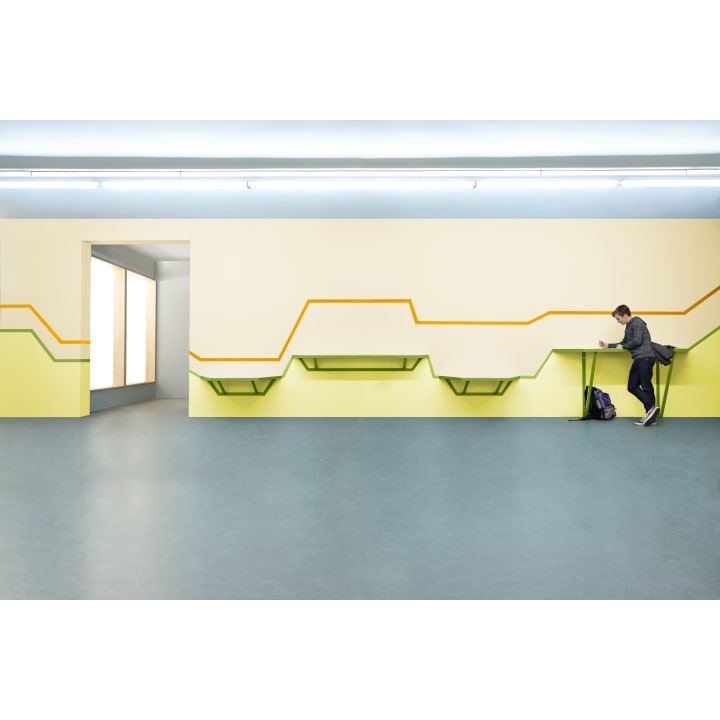 Eine Person steht an einem grünen Wandschreibtisch in einem minimalistischen Raum mit hellgelben Wänden, geometrischen Linien und blauem Bodenbelag. Es gibt mehrere unbesetzte Schreibtische und einen Rucksack auf dem Boden.