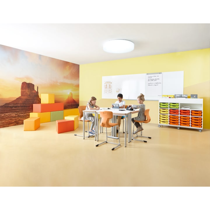 Drei Kinder sitzen an Tischen und arbeiten in einem hell erleuchteten Klassenzimmer mit gelben Wänden, bunten Ablagefächern, einer großen Wüstenlandschaft und stapelbaren Bauklötzen in einer Ecke.