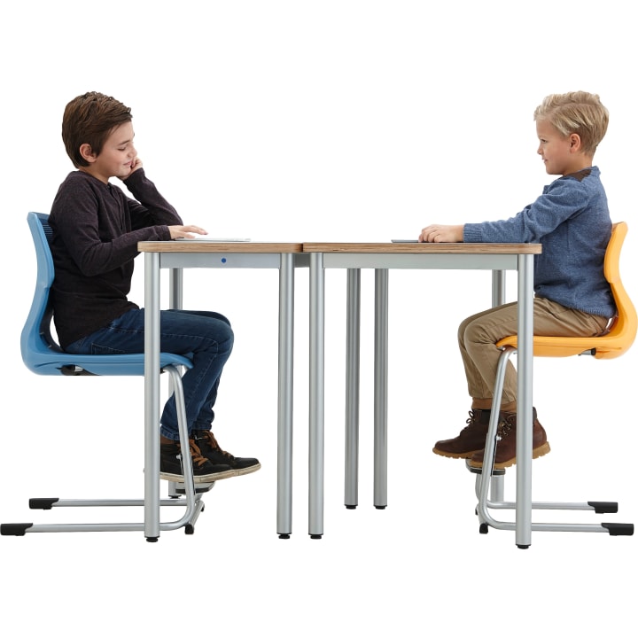 Zwei Jungen sitzen sich an einem Tisch mit Metallbeinen gegenüber. Der eine Junge sitzt auf einem blauen Stuhl und trägt eine Jeans und einen dunklen Pullover. Der andere Junge sitzt auf einem orangefarbenen Stuhl, trägt eine khakifarbene Hose und ein blaues Hemd. Beide haben Tablets auf dem Tisch liegen.