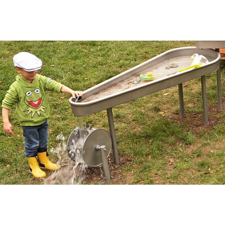 Ein kleines Kind in einem grünen Kermit-der-Frosch-Kapuzenpulli und gelben Regenstiefeln bedient einen Wasserspieltisch mit einer Kurbel, wodurch Wasser auf den Rasen fließt. Der Tisch hat schwimmende Spielzeuge auf seiner Oberfläche.