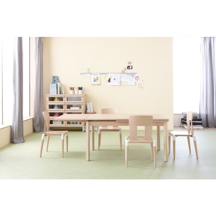 Ein Klassenzimmer mit vier Holzstühlen und einem rechteckigen Tisch, Regalen mit Büchern und Material und bunten Kinderzeichnungen an einer hellgelben Wand. Große Fenster lassen natürliches Licht herein.