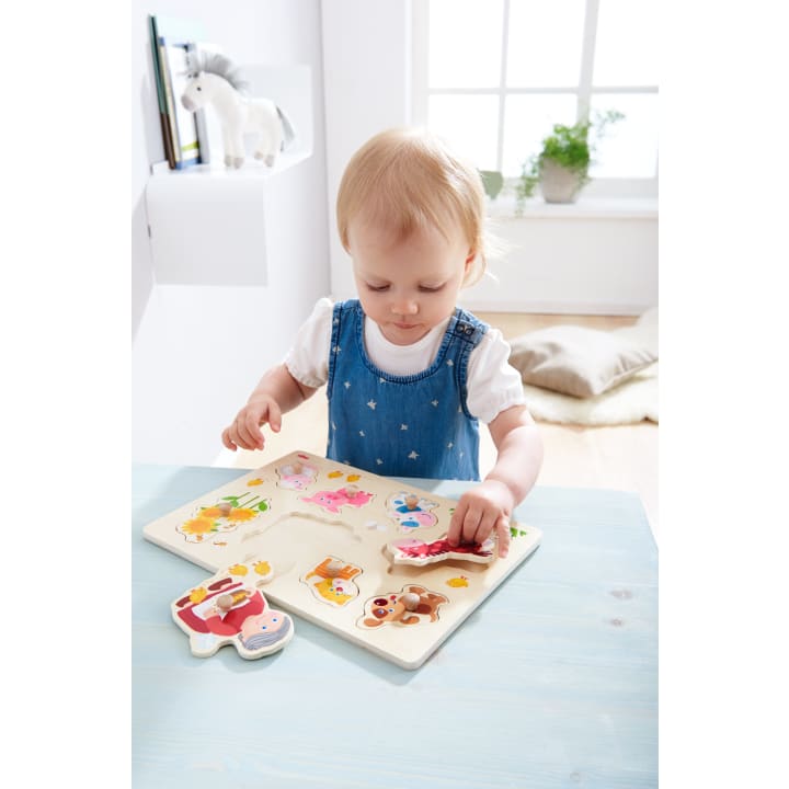 Ein kleines Kind in einem Jeanskleid legt ein hölzernes Tierpuzzleteil auf einen hellblauen Tisch in einem hellen Raum mit einem weichen Kissen, einer Pflanze und einem Spielzeugpferd auf einem Regal im Hintergrund.