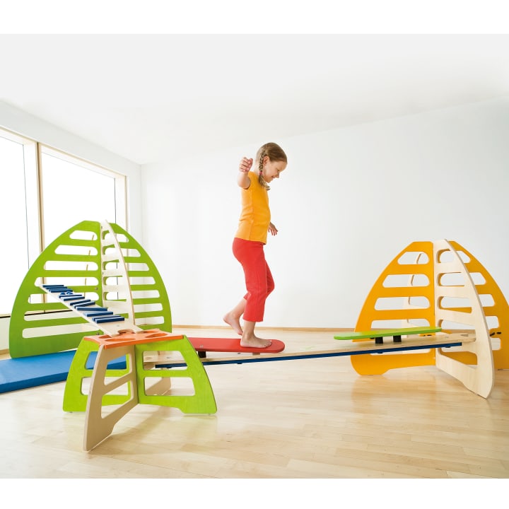 Ein Kind, das ein orangefarbenes Hemd und eine rote Hose trägt, balanciert auf einem hölzernen Spielgerät mit verschiedenen Rampen und Plattformen in einem hellen Raum mit Holzboden und großem Fenster.