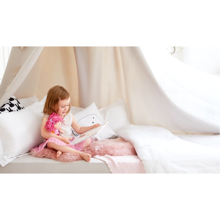 Ein junges Mädchen sitzt auf einem Bett unter einem weißen Zelt, hält eine Puppe und liest ein Buch. Das Bett hat weiße Kissen und eine rosa Decke, die eine gemütliche Atmosphäre schaffen.