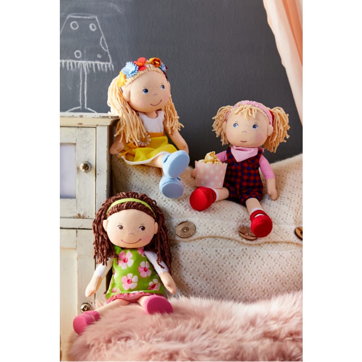 Drei Stoffpuppen mit Garnhaaren sitzen auf einer cremefarbenen Strickcouch; eine trägt ein gelbes Kleid mit einem Blumenstirnband, eine hat rote Schuhe mit Latzhose und eine Geschenktüte, und eine hat ein grünes Blumenkleid und ein Stirnband.