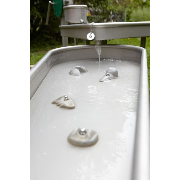 Nahaufnahme eines Wassertrogs mit Klettergriffen im Inneren, Wasser, das aus einem Ausguss oben in den Trog fließt, und Grünpflanzen im Hintergrund.