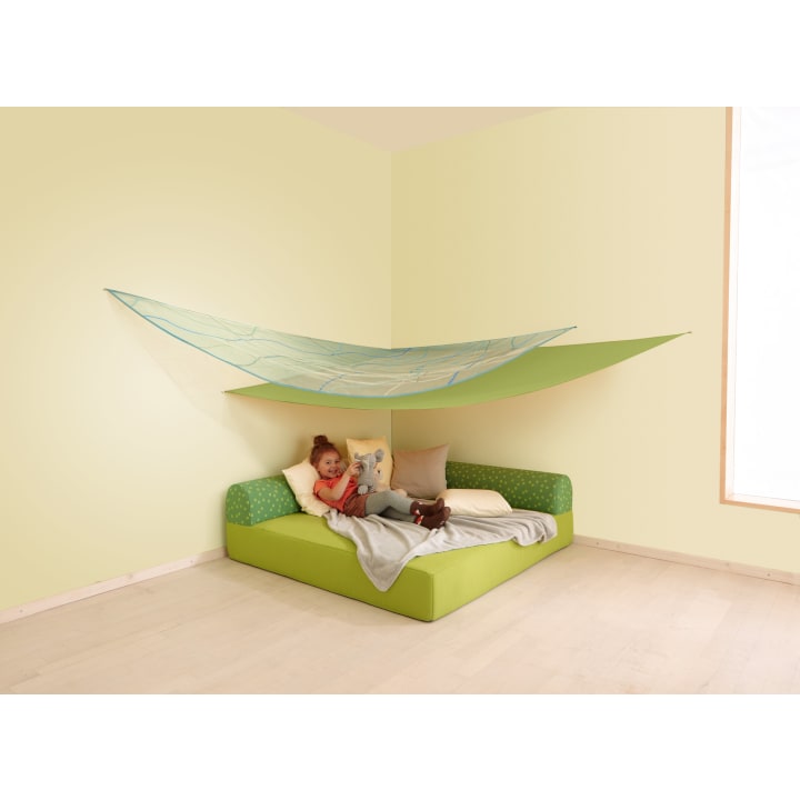 Ein Kind liegt auf einem grünen Bettsofa mit mehreren Kissen und einer grauen Decke und hält ein Stofftier. Über dem Sofa ist ein großer grüner Stoffhimmel angebracht, der einen zeltartigen Schutz in einem minimalistischen, hellen Raum schafft.
