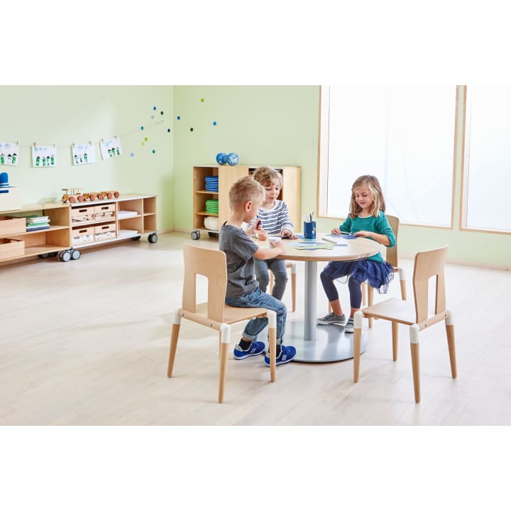 Drei kleine Kinder sitzen um einen runden Tisch in einem hellen Klassenzimmer und zeichnen oder schreiben auf Papier. Im Hintergrund sind Regale mit Material und Dekoration zu sehen. Der Raum hat helle Holzböden und große Fenster.