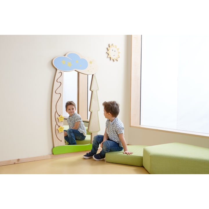 Ein kleiner Junge sitzt auf grünen Kissen in einem hellen Zimmer und betrachtet sein Spiegelbild in einem dekorativen Wandspiegel mit Wolken, Sternen und einem Baum. Über dem Spiegel hängt eine Sonnendekoration an der Wand.
