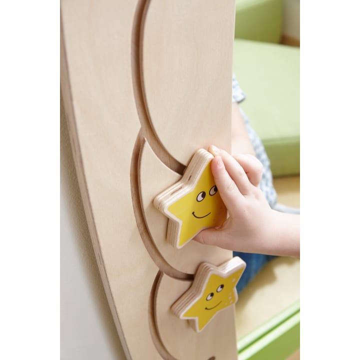 Die Hand eines Kindes bewegt einen gelben Holzstern mit einem lächelnden Gesicht entlang einer gebogenen Bahn auf einem hellen Holzbrett. Ein weiterer ähnlicher Stern ist darunter zu sehen. Im Hintergrund ist ein grünes Kissen zu sehen.