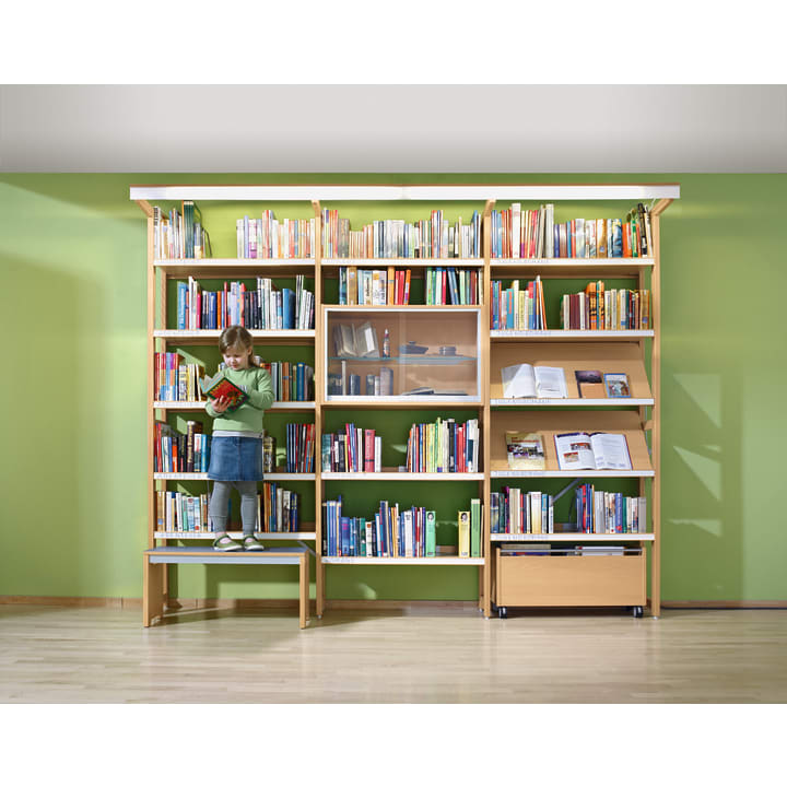 Ein kleines Kind steht auf einer Bank vor einem großen, mit bunten Büchern gefüllten Bücherregal und liest in einem roten Buch. Die Wand hinter dem Bücherregal ist hellgrün gestrichen. Der Boden ist aus hellem Holz.