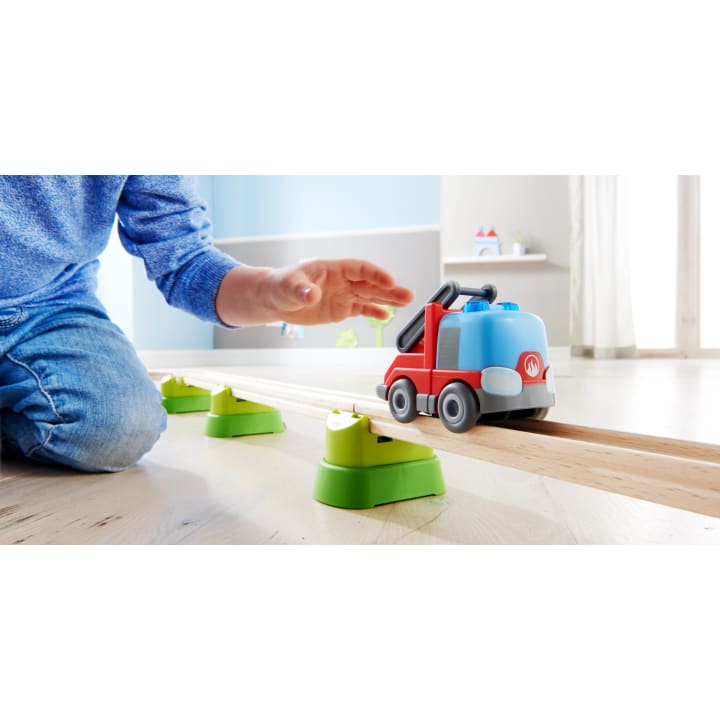 Ein Kind in einem blauen Pullover kniet auf dem Boden und spielt mit einem roten und blauen Spielzeug-LKW auf einer Holzschiene, die von grünen Ständern in einem hellen Raum getragen wird.