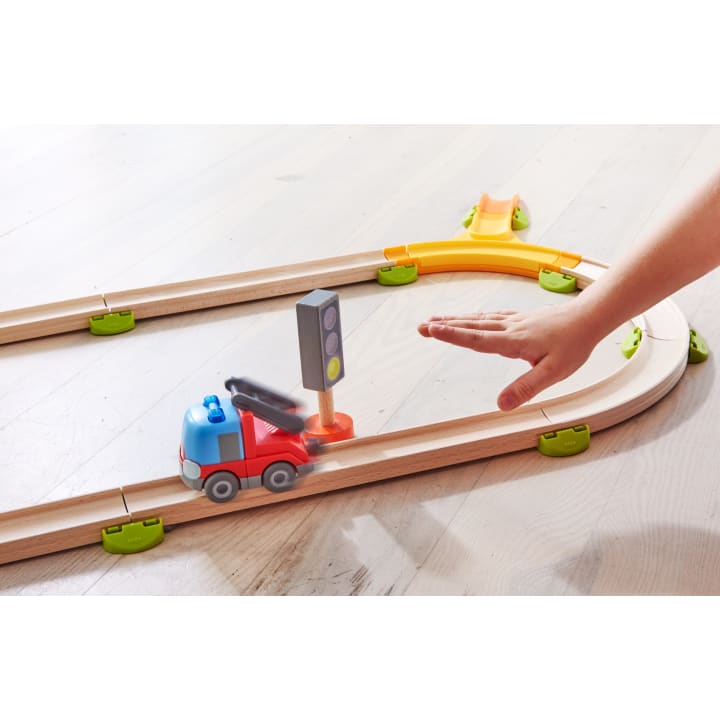 Eine Hand greift nach einem roten Spielzeug-Feuerwehrauto auf einer Holzeisenbahn, die mit einer Ampel, grünen Verbindungsstücken und einer gelben Rampe auf einem hellen Boden ausgestattet ist.